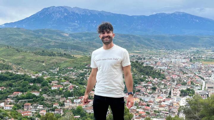 Stefan standing in front of a town and hills in the distance, wearing a white t-shirt that says 