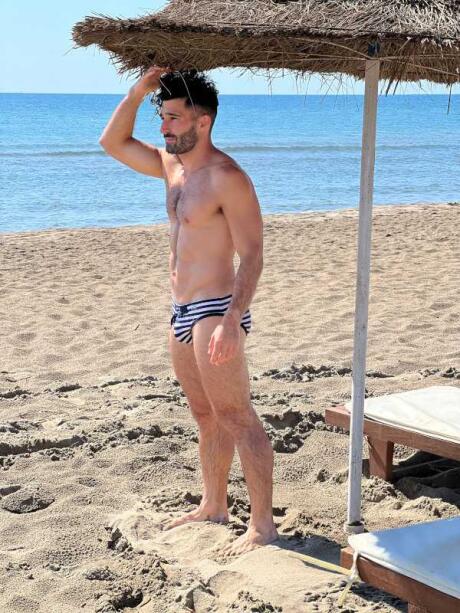 Stefan standing on a beach under a sun umbrella in a tiny speedo.