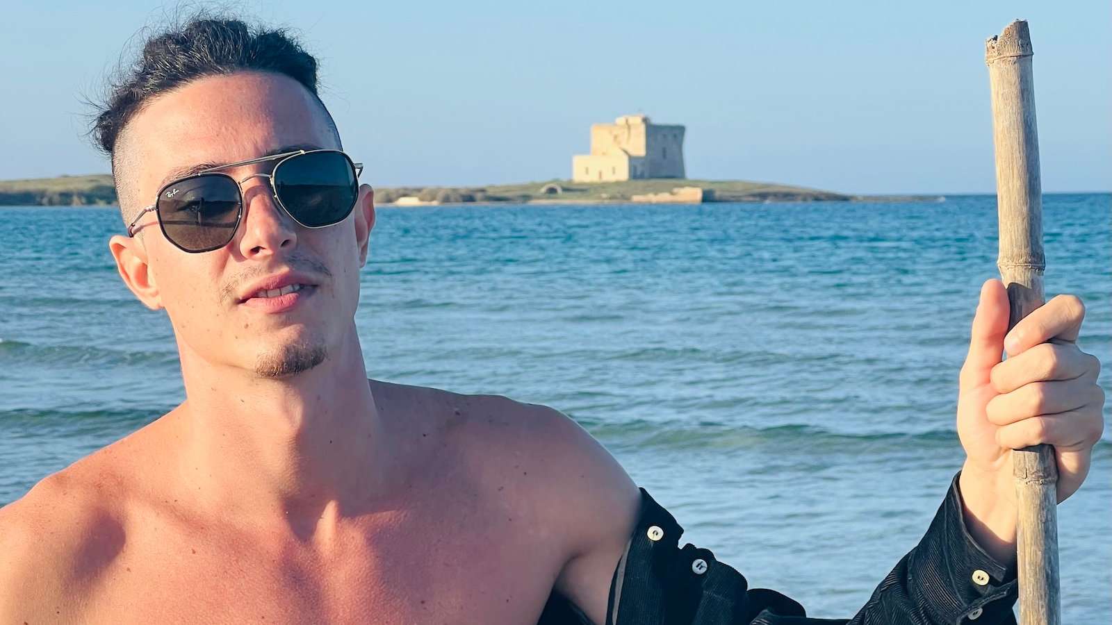 A man in sunglasses and his shoulders bare in front of a beach with a fortification in the distance.