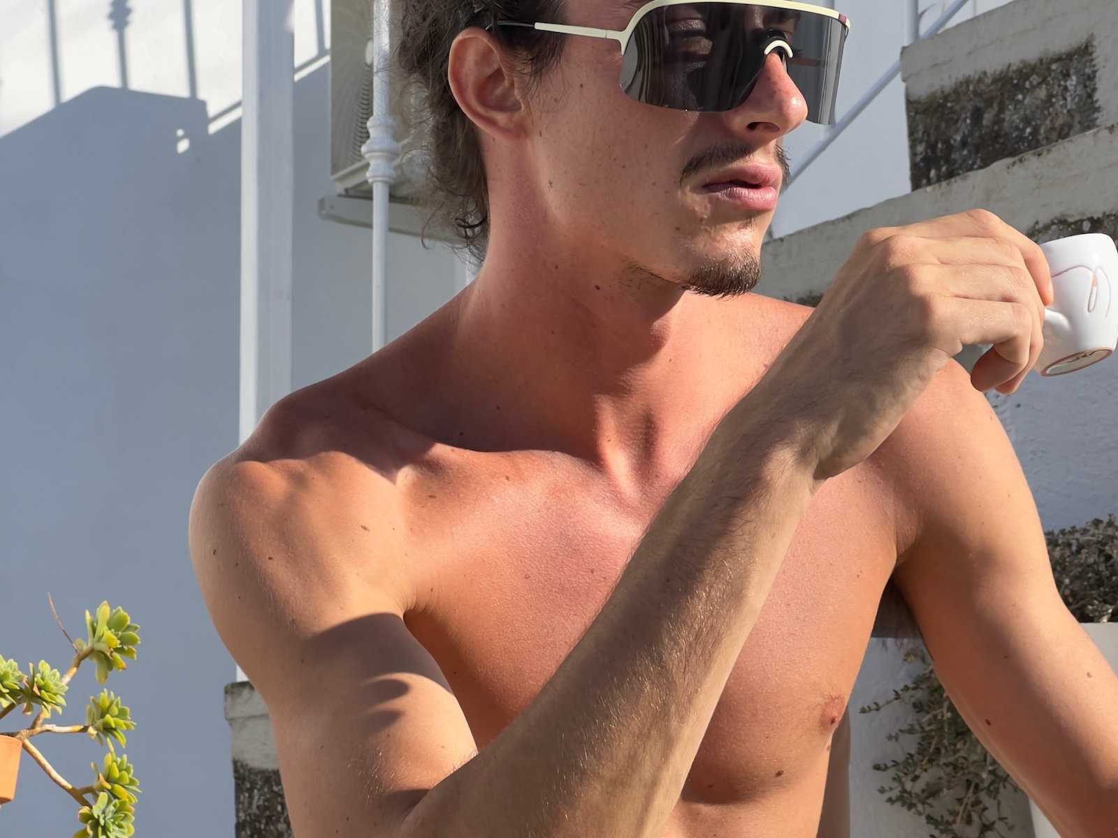 A close-up photo of a gay handsome man in Italy wearing sunglasses drinking coffee.